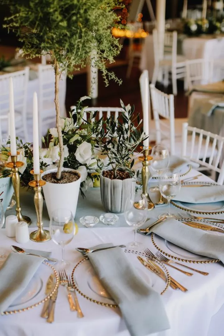 Круглый свадебный стол с золотыми приборами и живыми цветами в горшках.
