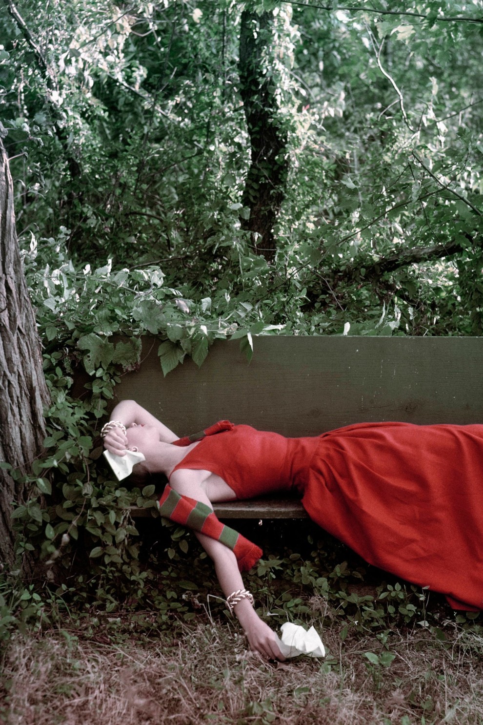 Модель лежит на скамейке в красном длинном платье с салфетками в руках.